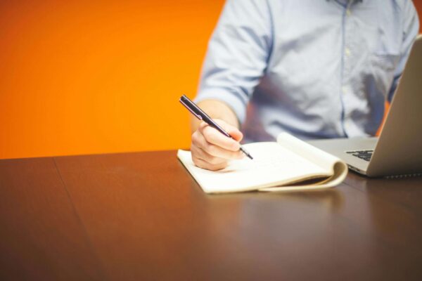 A person writing notes at a workspace with a laptop and vibrant orange background.