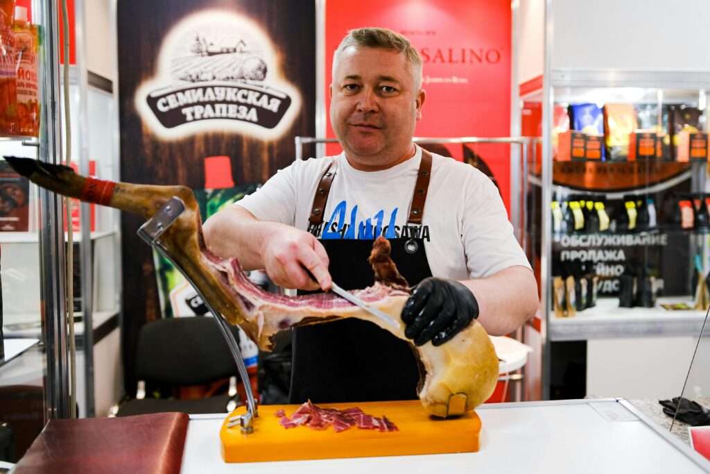 Man carving cured meat at a butcher shop exhibit indoors.
