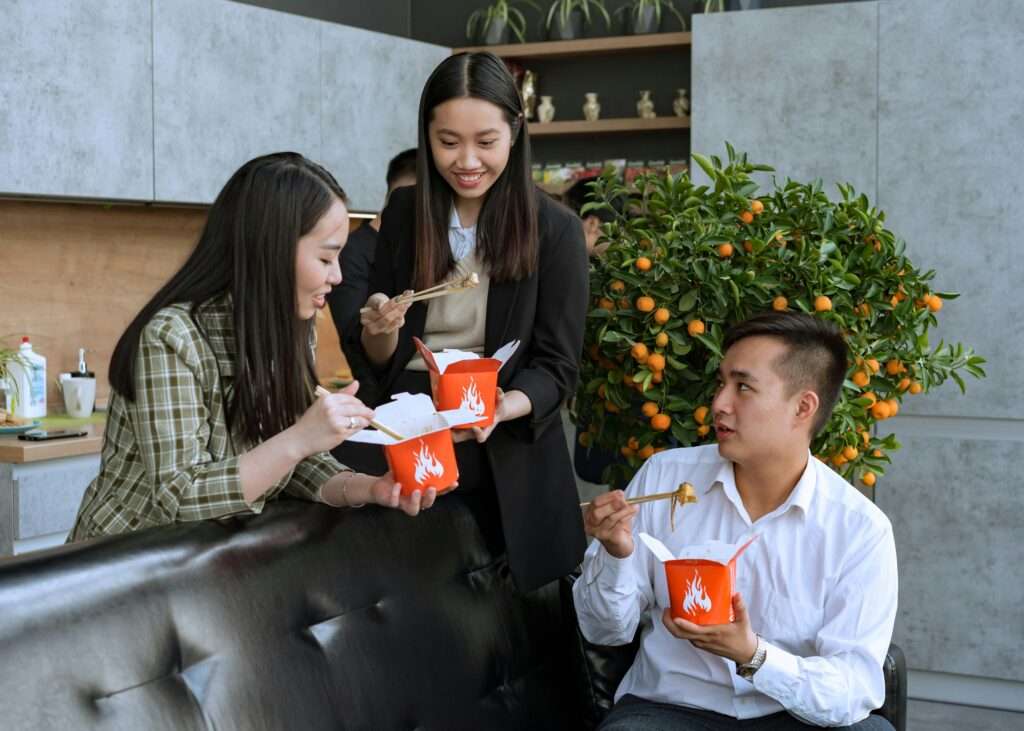 Group of colleagues enjoying a break eating takeout food with chopsticks indoors.